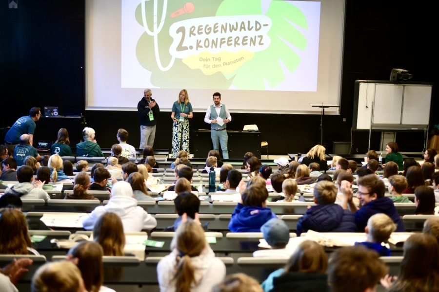 Vorlesesaal mit drei Personen vorne auf der Bühne, hinter ihnen eine Leinwand mit einem Bild. In den Sitzreihen sitzen viele Kinder mit dem Rücken zur Kamera.