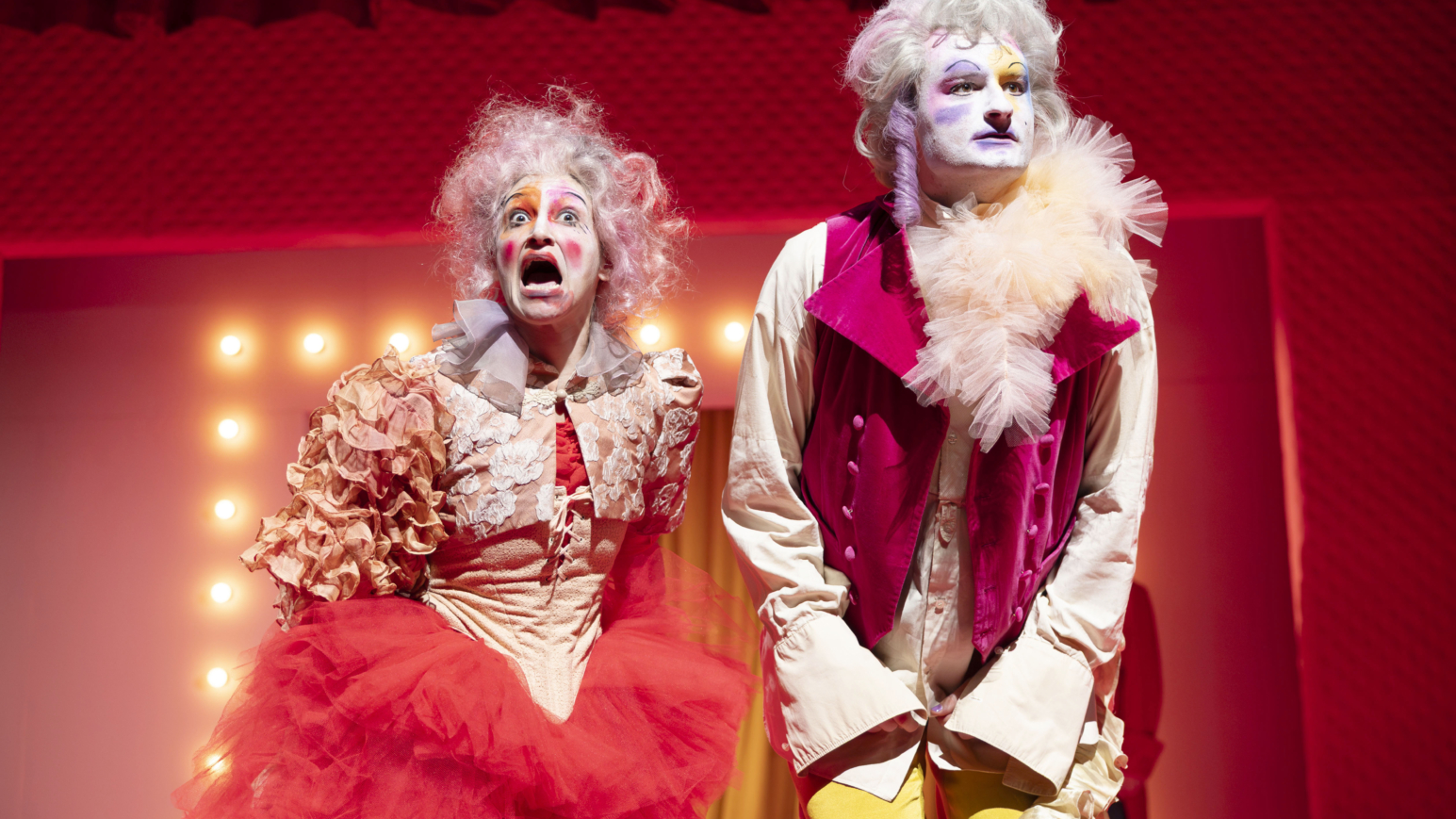 Zwei bunt gekleidete und geschminkte Theaterdarsteller:innen auf der Bühne.