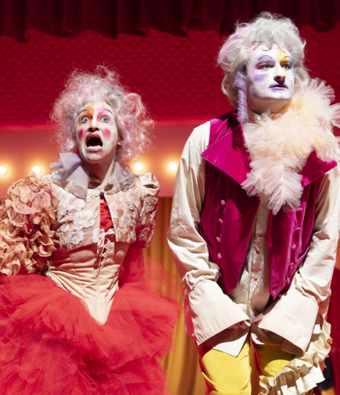 Zwei bunt gekleidete und geschminkte Theaterdarsteller:innen auf der Bühne.