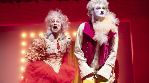 Zwei bunt gekleidete und geschminkte Theaterdarsteller:innen auf der Bühne.