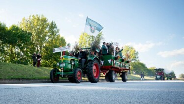 Grüner Traktor zieht einen Anhänger mit mehreren Menschen, die winken, auf einer Straße bei sonnigem Wetter und grünen Bäumen im Hintergrund.