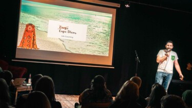 Ein Mann steht auf einer Bühne und spricht. Im Hintergrund ist eine Präsentation mit dem Titel 'Carpe Diem' und dem Untertitel 'Life Coaching' zu sehen. Der Mann trägt ein weißes T-Shirt und eine grüne Hose.