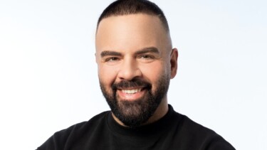 Mann mit kurzem, dunklem Haar und Bart trägt ein schwarzes Sweatshirt vor weißem Hintergrund.