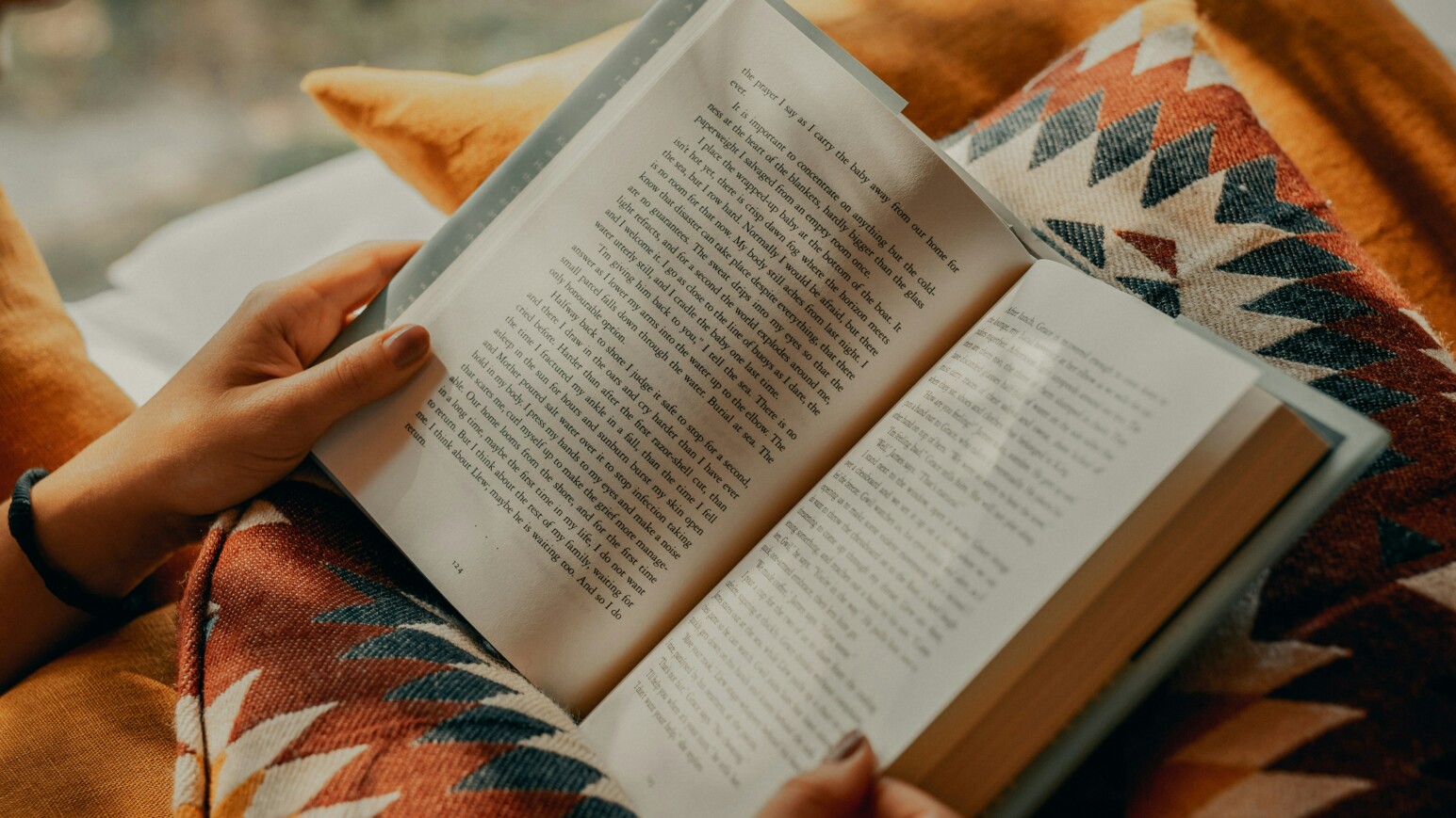 Hände halten ein aufgeschlagenes Buch mit Text, im Hintergrund eine Decke mit geometrischem Muster in Braun- und Orangetönen.