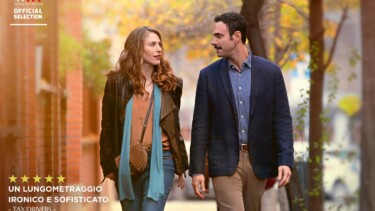 Filmposter mit zwei Personen, die nebeneinander auf einer Straße gehen. Links eine Frau mit langen, lockigen Haaren und brauner Jacke, rechts ein Mann in einem blauen Blazer.