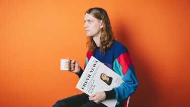 Daði Freyr sitzt auf einem Stuhl und hält eine Tasse in der linken Hand sowie eine Zeitung mit dem Titel 'FREYR NEWS' in der rechten Hand. Er trägt einen bunten Pullover mit roten, blauen und grünen Streifen.