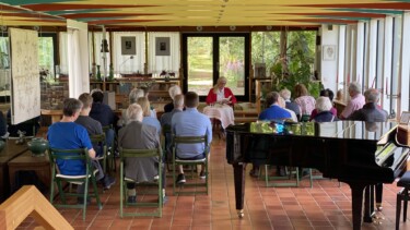 Mehrere Personen sitzen auf Stühlen in einem Raum mit großen Fenstern, ein Klavier steht rechts, vorne liest eine Person aus einem Buch vor.