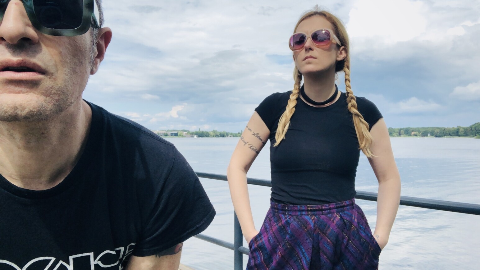 Eine Person mit Zöpfen trägt ein schwarzes T-Shirt und eine gemusterte Hose. Im Hintergrund ist ein Gewässer und bewölkter Himmel zu sehen.