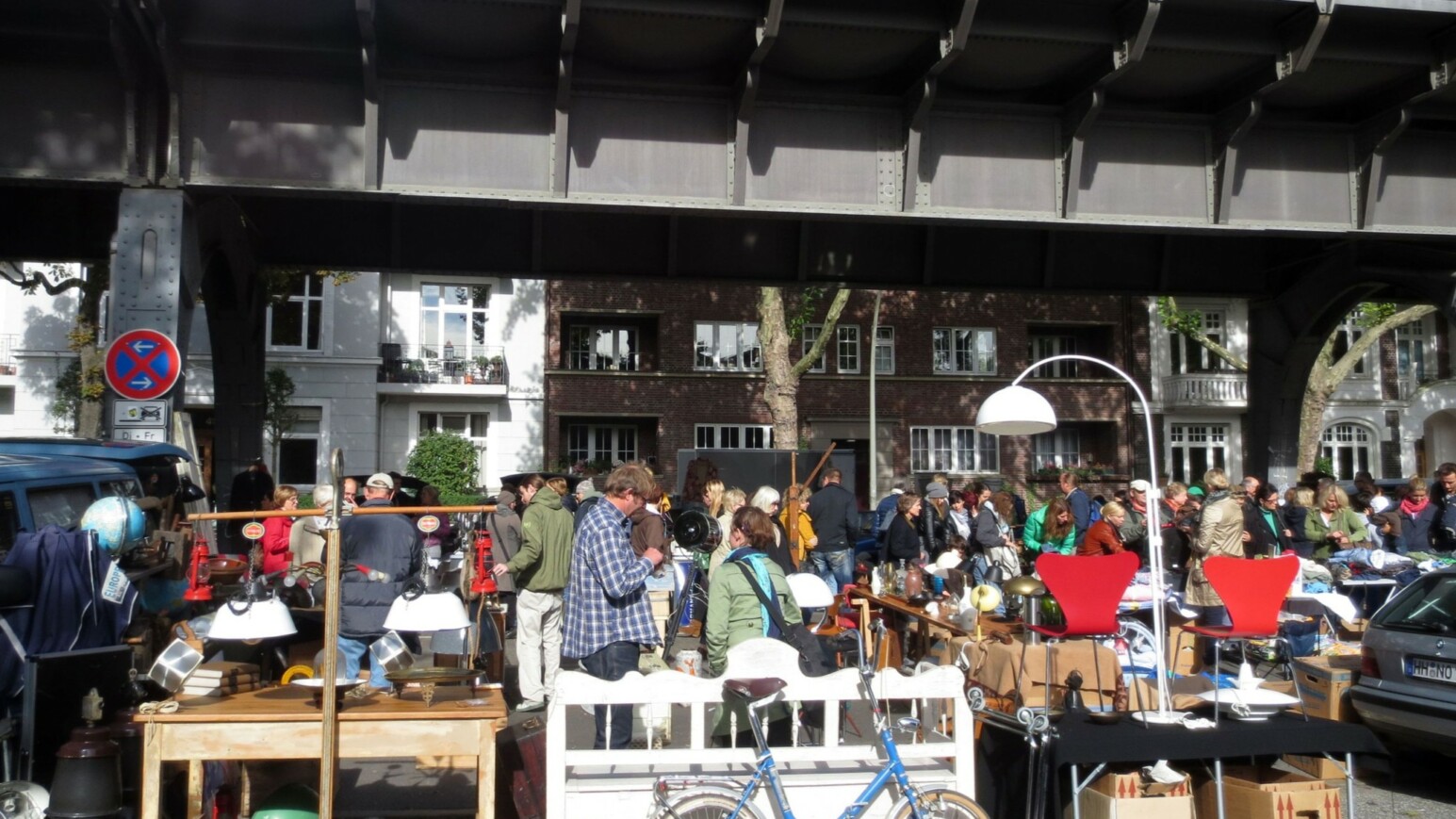 Menschenmenge auf einem Flohmarkt unter einer Brücke mit Tischen voller Lampen und anderen Gegenständen.