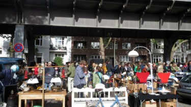 Menschenmenge auf einem Flohmarkt unter einer Brücke mit Tischen voller Lampen und anderen Gegenständen.