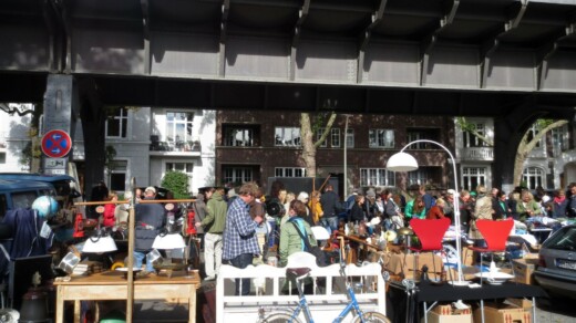 Menschenmenge auf einem Flohmarkt unter einer Brücke mit Tischen voller Lampen und anderen Gegenständen.