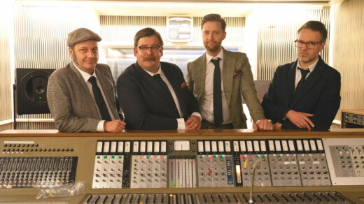 Vier Männer in Anzügen stehen hinter einem großen Mischpult in einem Tonstudio mit Lautsprechern und einem Radio im Hintergrund.