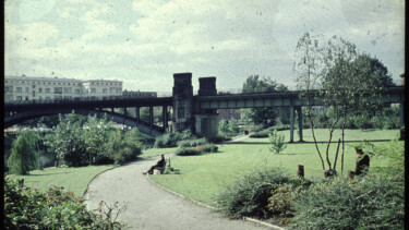 Grüner Park mit geschwungenem Weg, zwei Personen sitzen auf Bänken, im Hintergrund eine Eisenbahnbrücke und Wohngebäude.