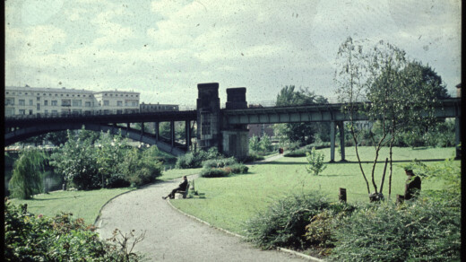 Grüner Park mit geschwungenem Weg, zwei Personen sitzen auf Bänken, im Hintergrund eine Eisenbahnbrücke und Wohngebäude.