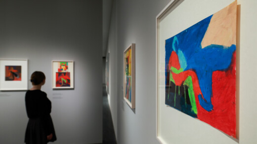 Eine Person mit einem schwarzen Kleid steht vor einem Kunstwerk mit bunten Formen in Blau, Rot und Gelb. Im Hintergrund sind weitere Bilder an der Wand.
