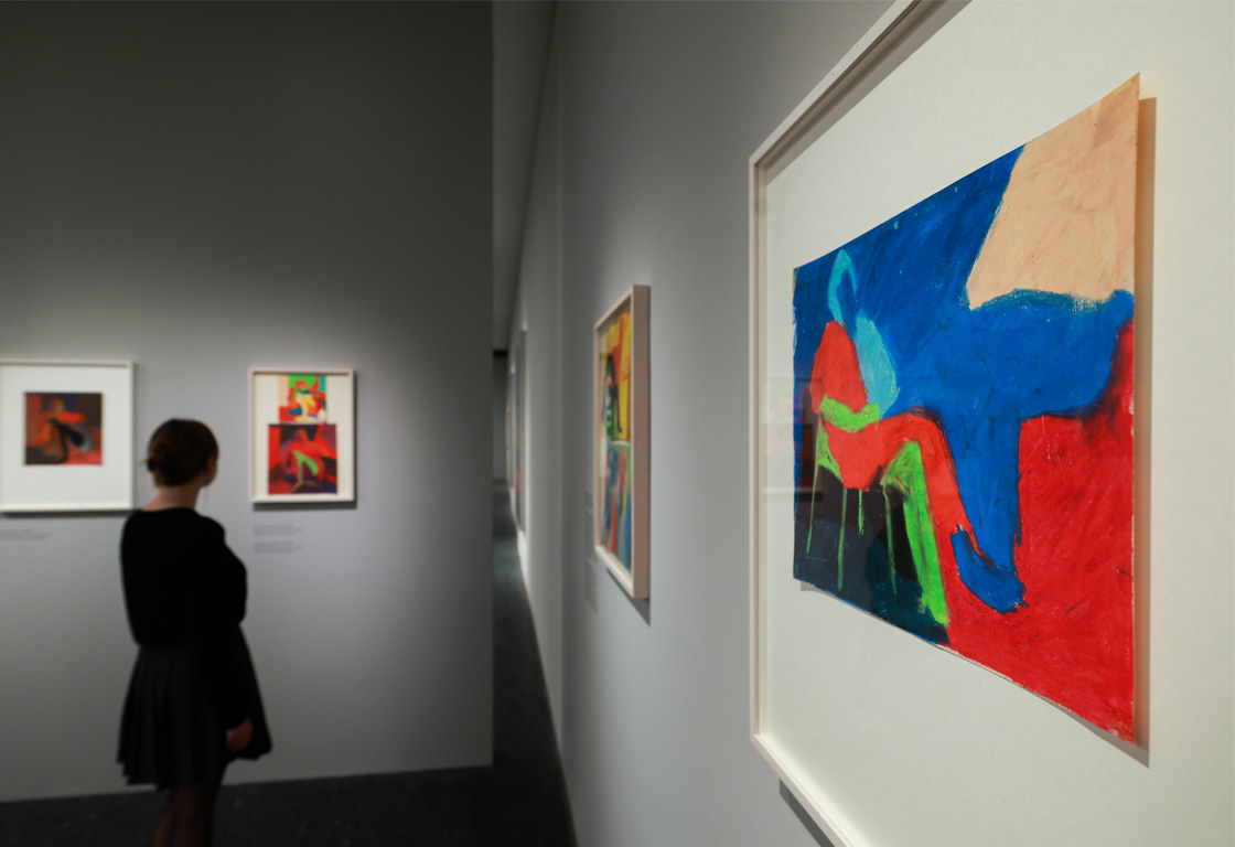 Eine Person mit einem schwarzen Kleid steht vor einem Kunstwerk mit bunten Formen in Blau, Rot und Gelb. Im Hintergrund sind weitere Bilder an der Wand.