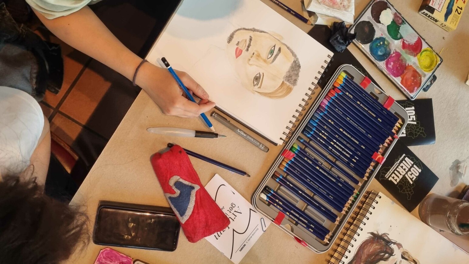 Hand zeichnet mit blauem Stift ein Porträt einer Frau mit roten Lippen auf einem Skizzenblock, daneben liegen Farbstifte, Aquarellfarben, ein Lineal, ein Radiergummi, ein rotes Etui und ein Smartphone.