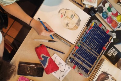 Hand zeichnet mit blauem Stift ein Porträt einer Frau mit roten Lippen auf einem Skizzenblock, daneben liegen Farbstifte, Aquarellfarben, ein Lineal, ein Radiergummi, ein rotes Etui und ein Smartphone.