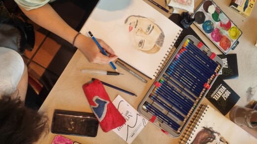 Hand zeichnet mit blauem Stift ein Porträt einer Frau mit roten Lippen auf einem Skizzenblock, daneben liegen Farbstifte, Aquarellfarben, ein Lineal, ein Radiergummi, ein rotes Etui und ein Smartphone.