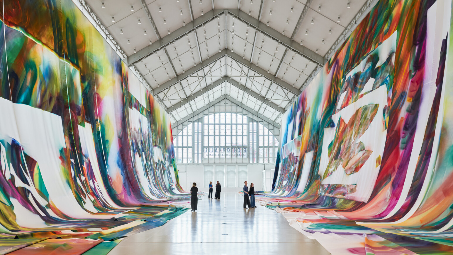 Große, farbenfrohe Wandmalerei in einer hellen Galerie mit hohen Decken. Menschen stehen und betrachten die Kunstwerke.