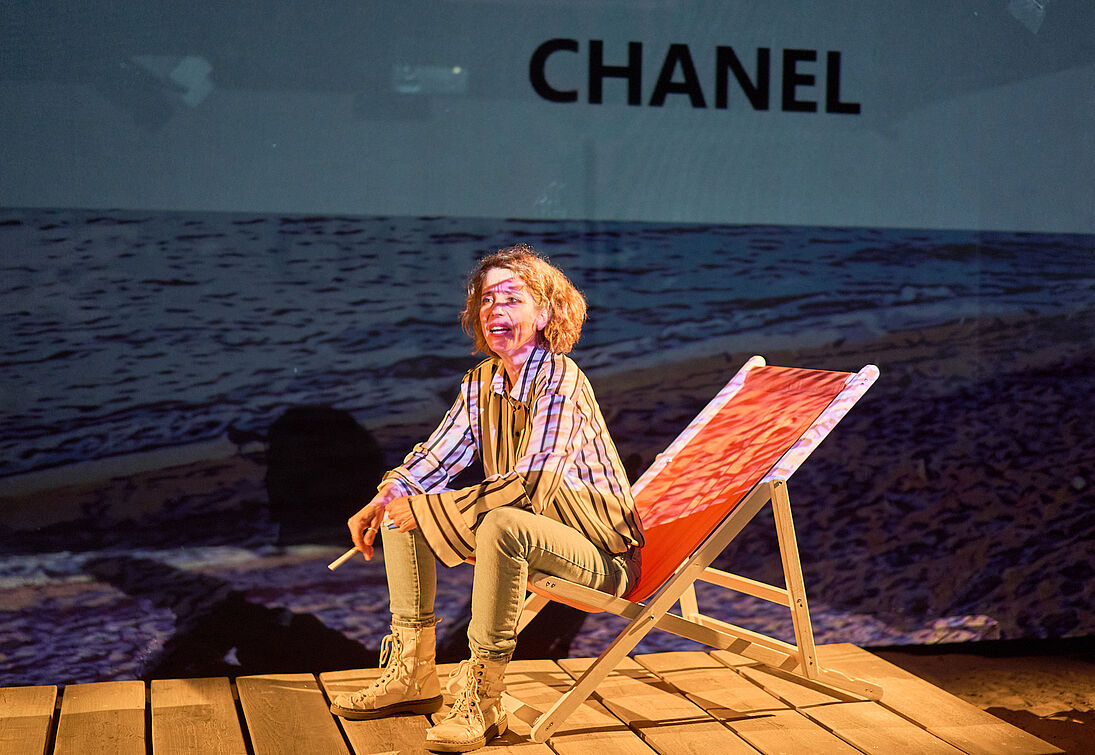 Bild von einer Frau in einem Liegestuhl auf einem Steg rauchend auf einer Bühne - Schrift Chanel