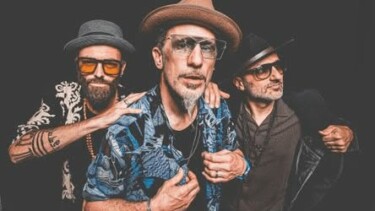 Die Band 'The Next Movement' mit vier Mitgliedern, die verschiedene Hüte tragen. Der Mann in der Mitte trägt ein blaues Hemd mit Mustern und eine Brille.