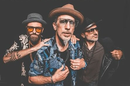 Die Band 'The Next Movement' mit vier Mitgliedern, die verschiedene Hüte tragen. Der Mann in der Mitte trägt ein blaues Hemd mit Mustern und eine Brille.