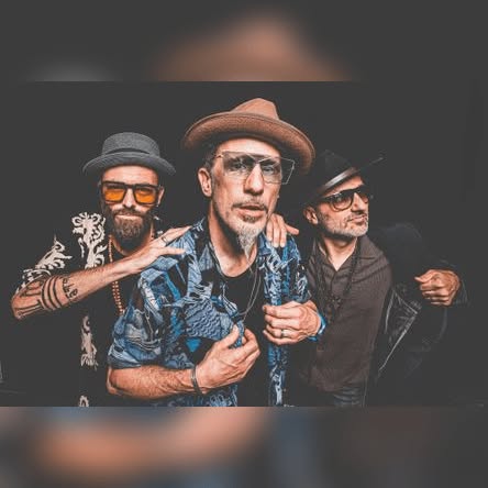 Die Band 'The Next Movement' mit vier Mitgliedern, die verschiedene Hüte tragen. Der Mann in der Mitte trägt ein blaues Hemd mit Mustern und eine Brille.