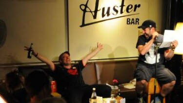 Bühne mit zwei Männern - Ein Mann sitzt auf einem Stuhl und liest vor, der andere sitzt dahinter auf einer Bank und hat ein Bier in der Hand