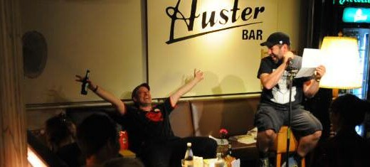 Bühne mit zwei Männern - Ein Mann sitzt auf einem Stuhl und liest vor, der andere sitzt dahinter auf einer Bank und hat ein Bier in der Hand