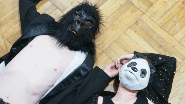 Zwei Personen liegen auf einem Holzboden, eine trägt eine schwarze Gorillamaske und ein schwarzes Jackett, die andere eine weiße Pandamaske und ein schwarzes, glitzerndes Oberteil.