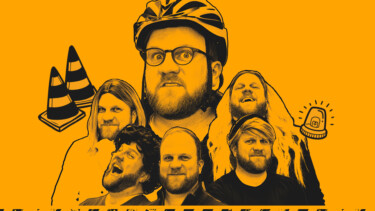 Fünf Personen vor orangefarbenem Hintergrund, eine trägt einen Fahrradhelm, zwei orange-weiße Verkehrskegel und ein blinkendes Warnlicht sind sichtbar.