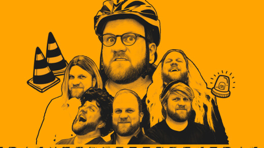 Fünf Personen vor orangefarbenem Hintergrund, eine trägt einen Fahrradhelm, zwei orange-weiße Verkehrskegel und ein blinkendes Warnlicht sind sichtbar.