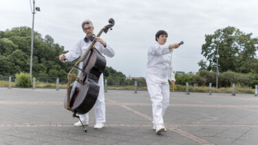 Zwei Personen in weißen Overalls, eine spielt Kontrabass mit Bogen, die andere hält ein Mikrofon und geht vorwärts auf einem gepflasterten Platz mit Bäumen im Hintergrund.