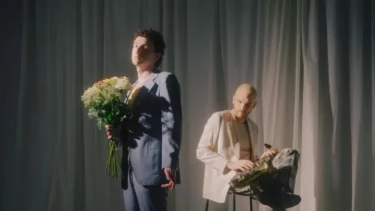 Zwei Personen vor einem Vorhang, eine steht mit einem Blumenstrauß in der Hand, die andere sitzt auf einem Hocker und hält ein Saxophon.