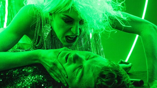 Person mit langen, lockigen Haaren und glitzerndem Kleid hält eine andere Person mit geschlossenen Augen am Hals, grüne Beleuchtung