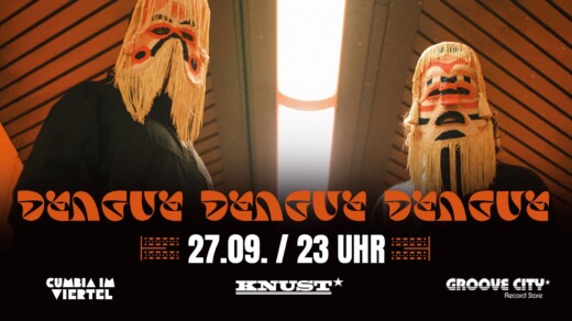 Zwei Personen mit langen, hellen Masken und schwarzen Kleidern vor einer Decke mit Leuchte.