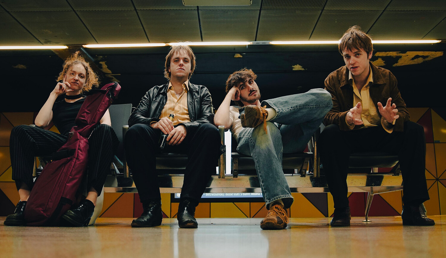 Vier Mitglieder der britischen Post-Punk-Band TALKIN‘ SECRET sitzen in einer Wartehalle. Zwei Personen tragen schwarze Kleidung, eine Person hat eine lila Tasche, die vierte Person trägt eine braune Jacke und Jeans.