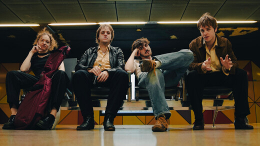 Vier Mitglieder der britischen Post-Punk-Band TALKIN‘ SECRET sitzen in einer Wartehalle. Zwei Personen tragen schwarze Kleidung, eine Person hat eine lila Tasche, die vierte Person trägt eine braune Jacke und Jeans.