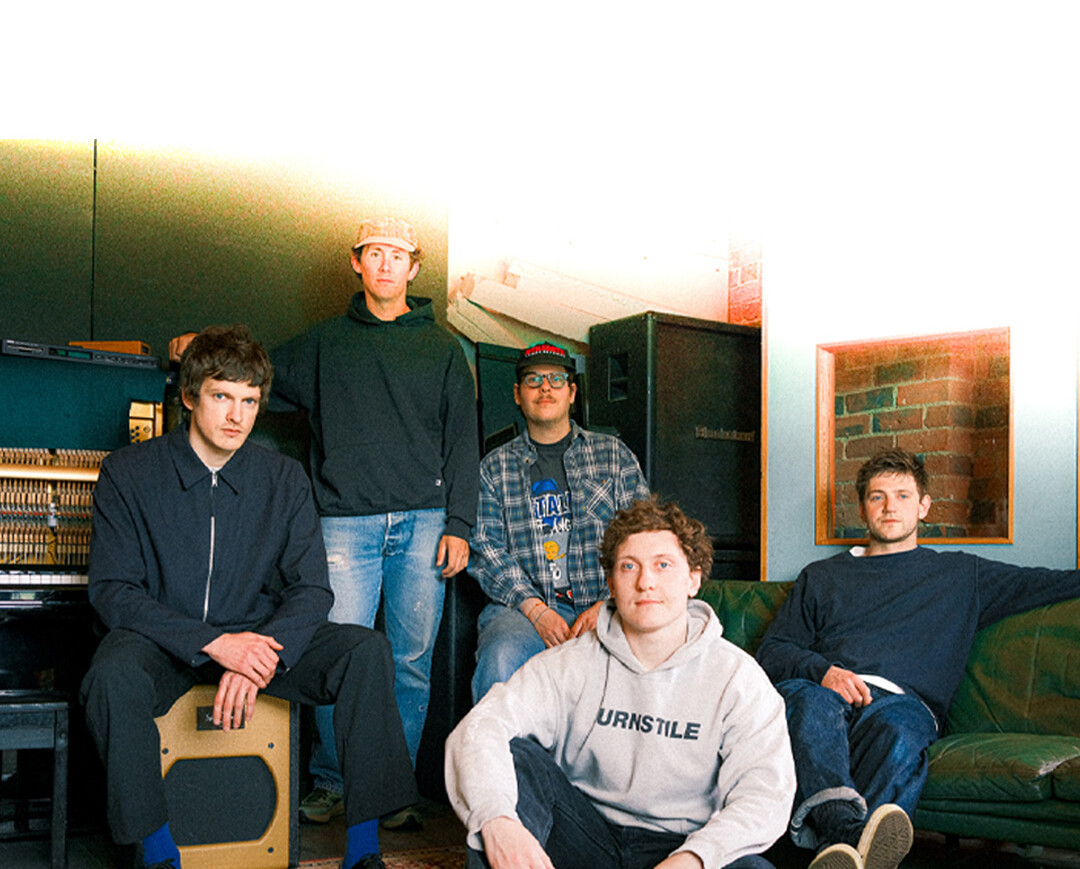 Die Band Basement steht in einem Raum. Fünf Mitglieder, einer sitzt auf einem Kasten, die anderen stehen oder sitzen auf einer Couch. Kleidung: lässig, mit Logos und Mustern.