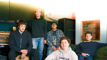 Die Band Basement steht in einem Raum. Fünf Mitglieder, einer sitzt auf einem Kasten, die anderen stehen oder sitzen auf einer Couch. Kleidung: lässig, mit Logos und Mustern.