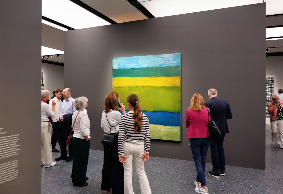Besucher stehen vor einem großen abstrakten Gemälde mit horizontalen Streifen in Blau, Grün und Gelb in einer Galerie.