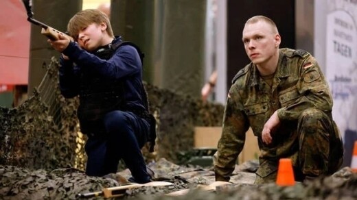 Ein Kind in militärischer Kleidung zielt mit einem Gewehr, während ein Soldat in Uniform neben ihm sitzt. Beide sind von militärischer Ausrüstung umgeben.