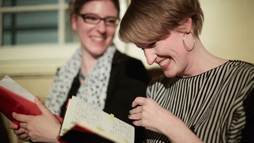 Zwei Personen sitzen nebeneinander, eine hält ein Notizbuch mit handschriftlichen Einträgen, die andere ein rotes Buch.