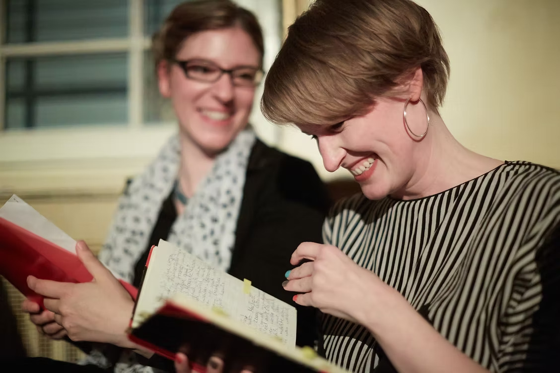 Zwei Personen sitzen nebeneinander, eine hält ein Notizbuch mit handschriftlichen Einträgen, die andere ein rotes Buch.
