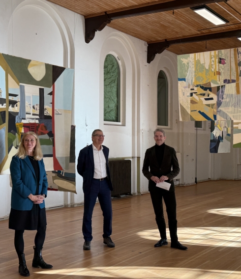 Auf dem Bild sind drei Personen in der Turnhalle der alten Schule, vor hängenden Gemälden zu sehen.