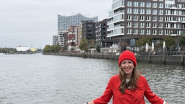 Citymanagerin in roter Kleidung lehnt an einer Reling; im Hintergrund die Elbe, Gebäude und die Elbphilharmonie.