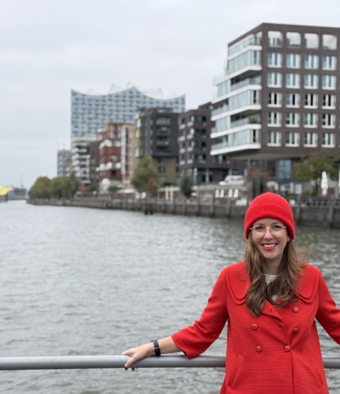 Citymanagerin in roter Kleidung lehnt an einer Reling; im Hintergrund die Elbe, Gebäude und die Elbphilharmonie.