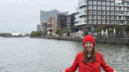 Citymanagerin in roter Kleidung lehnt an einer Reling; im Hintergrund die Elbe, Gebäude und die Elbphilharmonie.