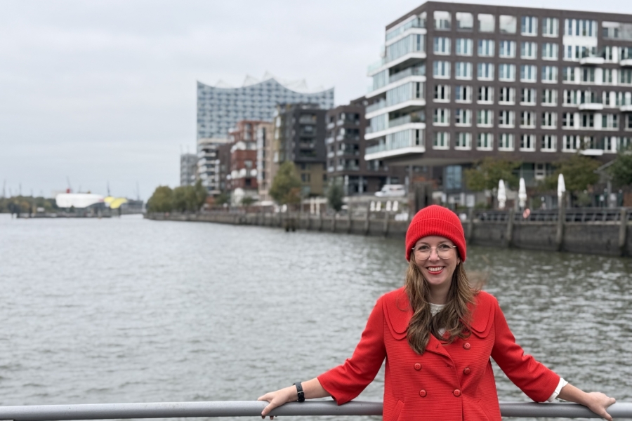 Citymanagerin in roter Kleidung lehnt an einer Reling; im Hintergrund die Elbe, Gebäude und die Elbphilharmonie.